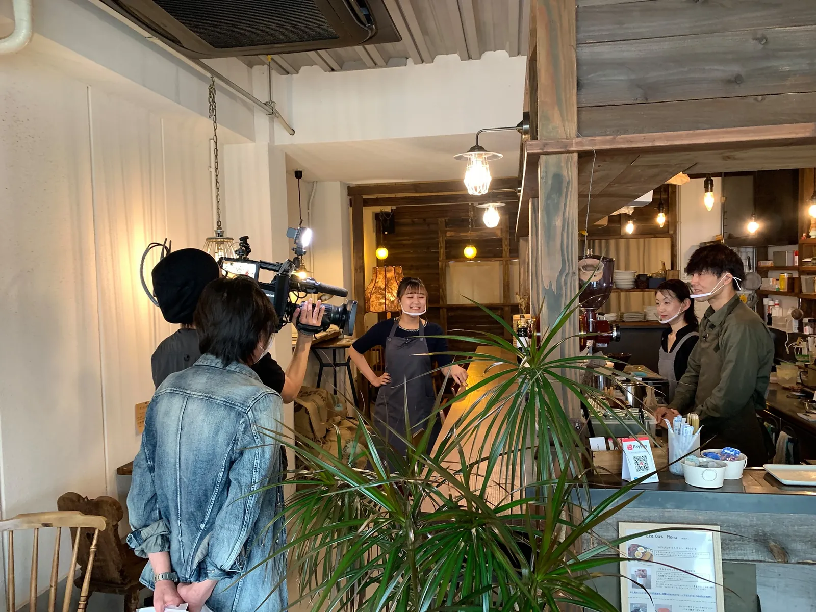 カフェの店内で、カメラを担いでスタッフを撮影している取材・動画制作の現場の様子。