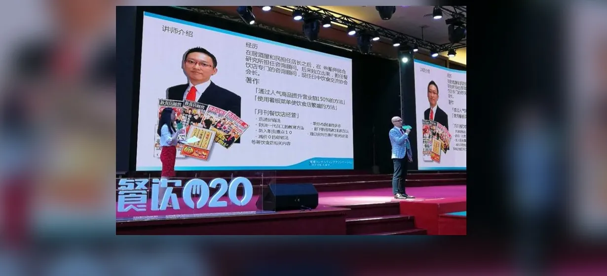 海外の業界カンファレンスでステージ中央に立ち、大型スクリーンに講師紹介スライドが映る中で話す宮内海。