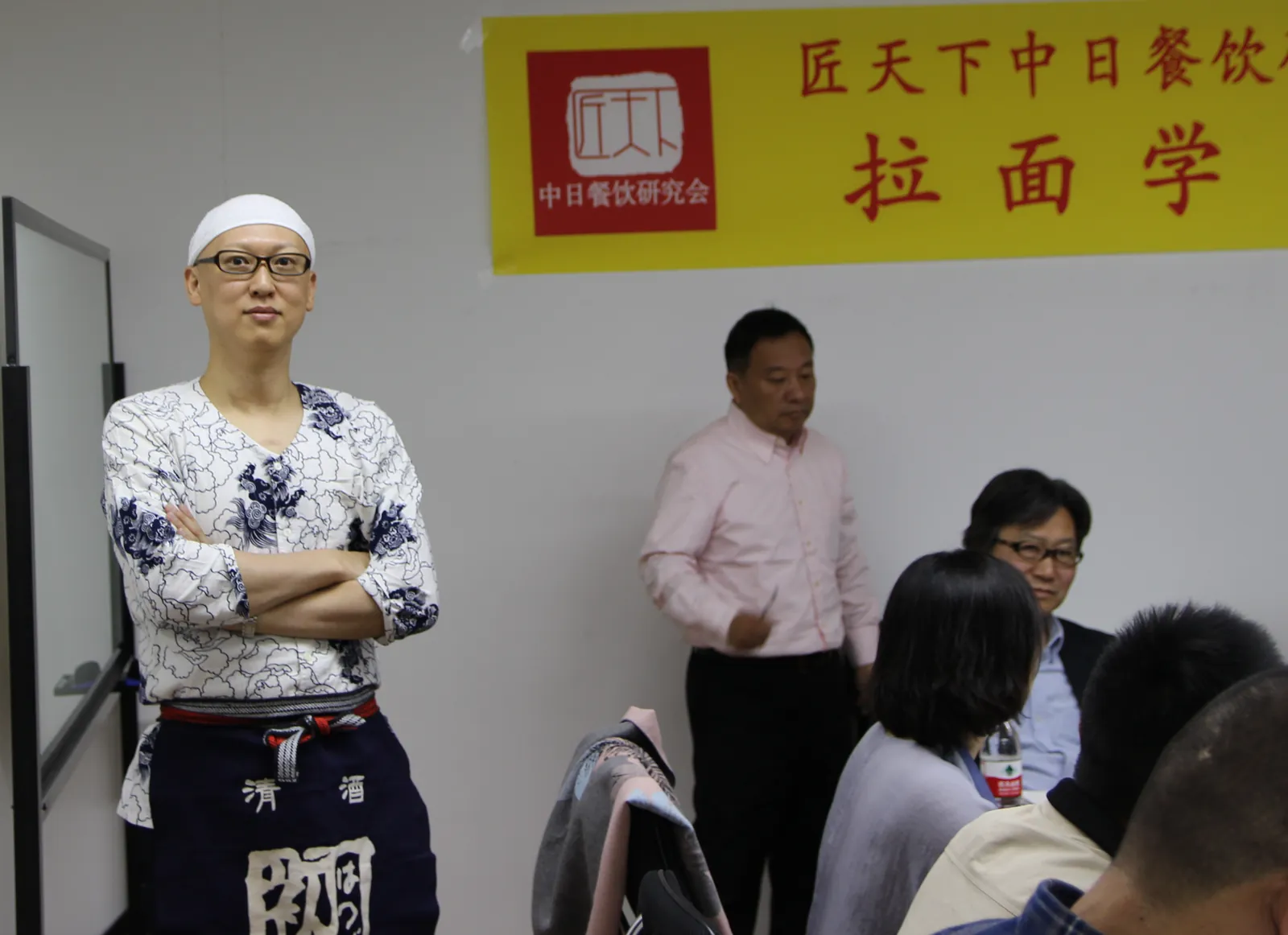 中国・匠天下中日餐飲研究会「拉面学院」の研修会場で、白い和柄の作務衣に前掛け姿で腕を組んで立つ宮内海。後方に参加者が着席している。