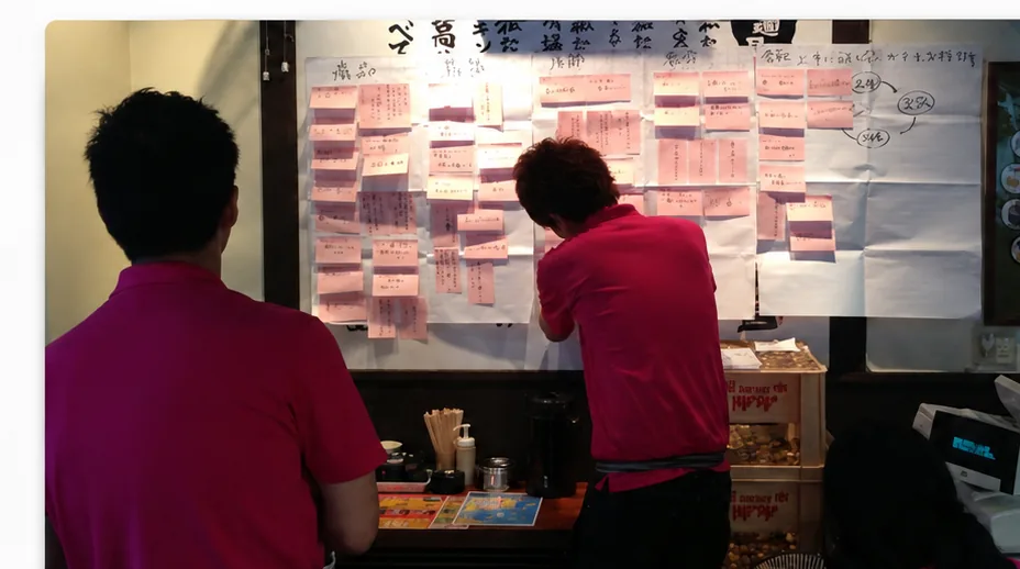 飲食店の現場で、付箋を貼り出したホワイトボードの前に立つスタッフ2名。営業会議で課題と改善点を出し合い、人が動く場づくりを実践している様子。